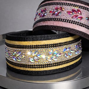 Op de afbeelding: Twee decoratieve hondenhalsbanden. De onderste halsband is geel met iriserende kristallen bladmotieven en zwarte kraalaccenten. De bovenste halsband is roze met vergelijkbare kristal- en kraaldetails. Beide halsbanden hebben zwarte randen.