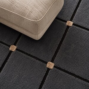 May include: A close-up of a dark grey rug with a geometric pattern of black lines and tan squares. A beige upholstered ottoman is partially visible in the upper left corner. The rug has a textured surface.