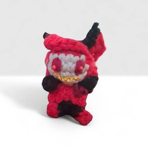 May include: A handmade crochet figure in red, black, and gray. The figure has a smiling face with yellow teeth, red eyes, and black accents. The figure is standing upright on a white surface.