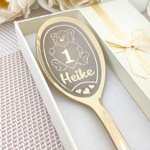 Peut inclure: Une cuillère dorée avec un motif gravé. Le motif comprend un ours en peluche tenant un cœur avec le chiffre 1, le nom "Heike" et des cœurs. La cuillère est dans un coffret cadeau blanc avec un ruban.
