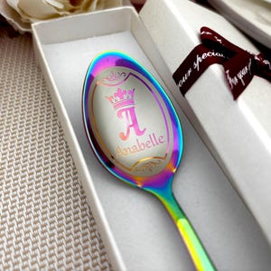 May include: A personalised spoon with a rainbow finish, presented in a gift box. The spoon features a crown design, the letter "A", and the name "Anabelle" engraved on the spoon's head.