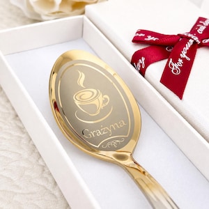 Könnte beinhalten: Ein goldfarbener Metalllöffel mit einer Gravur einer dampfenden Kaffeetasse und dem Namen Grażyna. Der Löffel liegt in einer weißen, rechteckigen Geschenkbox, die mit einem dunkelroten Band mit weißem Schreibschrifttext verziert ist.