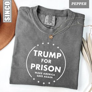 以下が含まれることがあります： 白の「TRUMP FOR PRISON」の文字が円で囲まれ、その下に「MAKE AMERICA SAFE AGAIN」と書かれた、ペッパーカラーのTシャツ。クルーネックと半袖です。ペンダント付きのシルバーネックレスがシャツの上にかけられています。