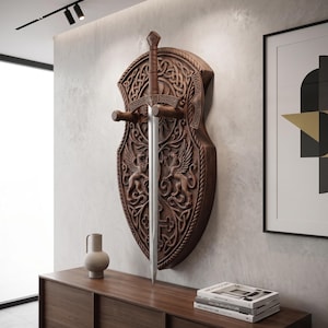Könnte beinhalten: Ein an der Wand montierter Holzschild mit aufwendigen Schnitzereien von zwei geflügelten Löwen und keltischen Knotenmustern. Ein langes, gerades Schwert mit braunem Ledergriff ist vertikal angebracht und wird von zwei horizontalen Holzstiften gehalten.