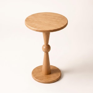 Puede incluir: Una mesa auxiliar de madera de color marrón claro con una superficie circular, una base central con un detalle esférico y una base redonda a juego. La veta natural de la madera es visible en toda la superficie lisa y pulida del mueble.