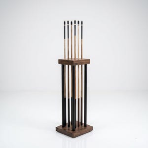 May include: A wooden pool cue rack holding six cues. The rack is made of dark brown wood with a square base and top, and vertical supports. The cues are a mix of light and dark wood, with black and white tips.