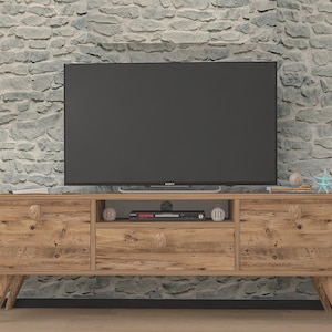 May include: A mid-century modern style TV stand made of light brown wood. The stand has two doors, a drawer, and open shelving for media components. The stand is supported by angled legs and has a large black television on top.