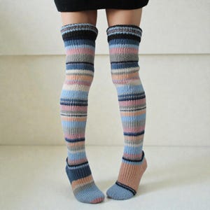 Puede incluir: Un par de calcetines de punto hasta el muslo con un patrón de rayas horizontales multicolor en tonos azul, rosa, gris y marrón. Los calcetines tienen una textura acanalada, una banda marrón oscuro en la parte superior y un talón reforzado.