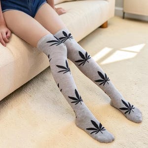 Può includere: Calzini alti grigi al ginocchio con un motivo ripetuto di foglie di cannabis nere. I calzini sono realizzati in un materiale morbido e sono adatti per l'abbigliamento casual. I calzini sono indossati da una persona seduta su un divano beige.