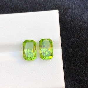 Puede incluir: Dos gemas rectangulares de color verde claro con una apariencia vibrante y translúcida. Las gemas están colocadas sobre una superficie blanca, mostrando su claridad y color. Las piedras son probablemente peridotos, conocidos por su tono verde oliva.