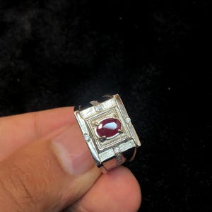Peut inclure: Bague en argent avec un design rectangulaire orné d'une pierre précieuse ovale rouge foncé au centre. De petites pierres claires sont serties autour de la pierre centrale. La bague est présentée sur fond sombre.