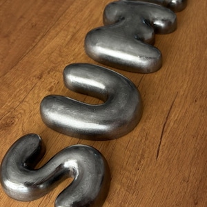May include: Four individual metallic, silver-colored, three-dimensional letters arranged vertically to spell the word "Suns". Each letter features a smooth, rounded, bubble-like shape with a polished finish that reflects light across its curved surfaces.