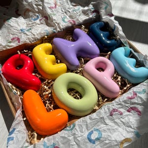 May include: A collection of seven glossy, rounded, three-dimensional ceramic letters in vibrant colors including red, yellow, orange, light green, purple, pink, and blue, nestled inside a cardboard box lined with patterned tissue paper.