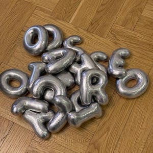 May include: A collection of individual silver-coloured metal letters with a brushed finish. The letters are shaped like rounded, inflated balloons and are scattered together in a pile, featuring various characters including O, S, H, R, and Y.
