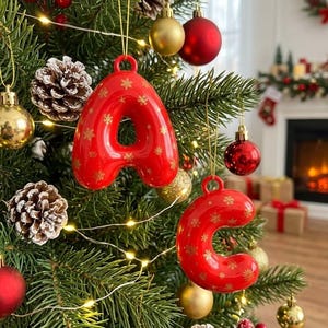 May include: Two glossy red Christmas ornaments shaped like the letters A and C, featuring a repeating gold snowflake pattern. Each letter ornament has a small loop at the top for hanging and is suspended by a thin gold string on a pine tree.