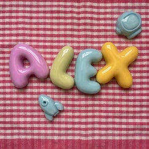Peut inclure: Un ensemble de quatre lettres brillantes, arrondies et aux couleurs pastel formant le nom ALEX, avec un A violet clair, un L jaune pâle, un E bleu clair et un X jaune vif, accompagné d'une petite fusée bleu clair et d'un casque bleu clair.