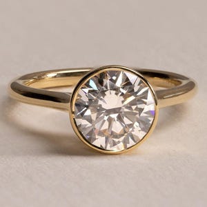 May include: A gold ring featuring a single round brilliant-cut clear gemstone set in a smooth bezel setting. The band is polished yellow gold with a rounded profile, showcasing a clean and simple design that highlights the central stone.
