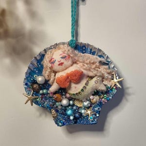 May include: A hanging ornament featuring a needle-felted mermaid with blonde hair and an orange top, nestled inside a blue shell decorated with various beads, pearls, and small gold-toned starfish. A teal yarn loop is attached for hanging.