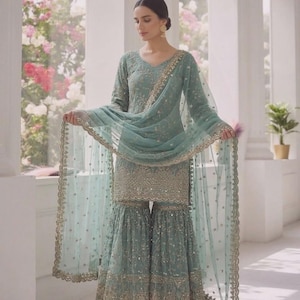 May include: A light teal Sharara suit with intricate gold embroidery. The ensemble includes a long-sleeved tunic, wide-legged pants, and a sheer dupatta. The outfit is embellished with sequins and delicate detailing, creating an elegant look.
