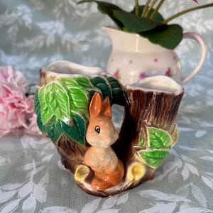 Könnte beinhalten: Ein Keramik-Pflanzgefäß in Form eines Baumstumpfes mit einer kleinen Hasenfigur. Der Pflanztopf hat zwei Öffnungen für Pflanzen, mit grünen Blättern und braunen Rinden Details. Der Hase ist orange und weiß und verleiht der Dekoration einen skurrilen Touch.