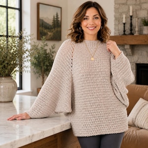 May include: A beige, loose-fitting crochet sweater with wide sleeves. The sweater has a round neckline and is worn with a gold pendant necklace. The model is standing in a well-lit room with a neutral color palette.
