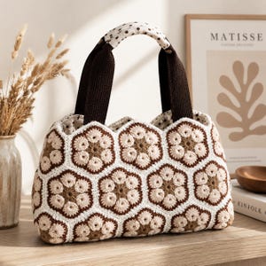 Puede incluir: Bolso de mano de ganchillo hecho a mano con un estampado floral en tonos marrón, crema y blanco. El bolso presenta un diseño hexagonal, un asa de lunares y correas marrones oscuras. El bolso está sobre una superficie de madera.