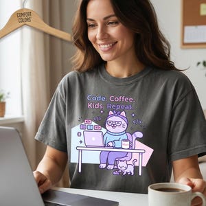 May include: A charcoal gray t-shirt with the text "Code. Coffee. Kids. Repeat." and a cartoon cat graphic. The cat is wearing glasses and a headband, sitting at a desk with a laptop. A coffee mug is on the desk. The shirt is hanging on a wooden hanger.