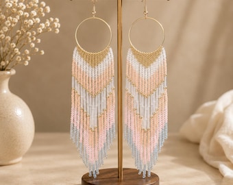 May include: A pair of long dangle earrings featuring gold-tone circular hoops at the top, with cascading fringe made of multi-colored seed beads in shades of gold, white, soft pink, and light gray, arranged in a V-shaped chevron pattern.