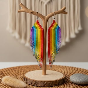 May include: A pair of vibrant, handmade beaded earrings in a rainbow color scheme. The earrings are displayed on a wooden stand, showcasing their intricate design and the array of colors, including red, orange, yellow, green, blue, and purple.
