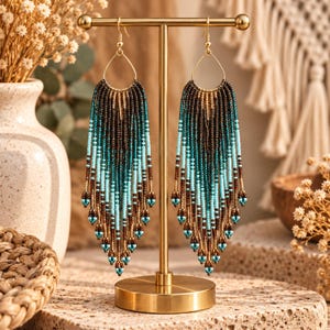 May include: A pair of long, teardrop-shaped dangle earrings featuring a gradient of seed beads in shades of dark brown, teal, and light blue. The earrings are suspended from gold-toned metal hooks and hang from a gold-toned metal T-bar display stand.