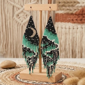 May include: Beaded dangle earrings in a celestial design. The earrings feature a black background with a crescent moon and turquoise, white, and black accents. The earrings are displayed on a wooden stand.