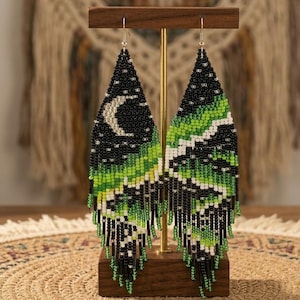 May include: Handmade beaded earrings in a triangular shape. The earrings feature a black background with a crescent moon design and green and white accents. The bottom of the earrings have long black and green fringe.