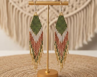 May include: A pair of beaded earrings in a teardrop shape, displayed on a gold-colored stand. The earrings feature a pattern of green, white, and rust-colored beads, with gold-colored beads at the bottom. The background includes a woven wall hanging.