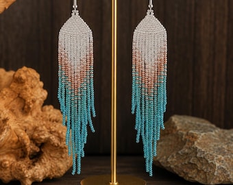 May include: A pair of long fringe earrings featuring a gradient pattern of small beads. The color transitions from a metallic silver at the top to a soft copper in the middle, ending in a vibrant turquoise blue at the bottom of the dangling strands.