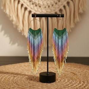 May include: A pair of beaded earrings with a gold hoop and a long fringe. The top is green, transitioning to blue, purple, pink, and yellow. The fringe ends with white and green beads. The earrings are displayed on a black stand.