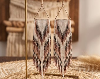 May include: A pair of beaded earrings with a geometric design. The earrings have a triangular top and a fringe of white, brown, and green beads. They are displayed on a gold-coloured stand.