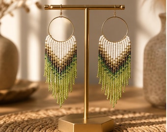 May include: A pair of beaded fringe earrings hanging from a gold-toned T-bar display stand. The earrings feature a circular gold hoop top with a gradient pattern of white, dark brown, and light green seed beads arranged in a V-shaped fringe.