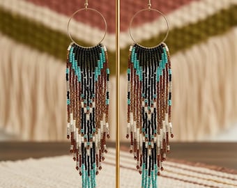 May include: A pair of long, beaded earrings with a gold-tone hoop and fringe. The earrings feature a pattern of black, turquoise, brown, and white beads. The earrings are displayed on a gold earring stand against a neutral background.