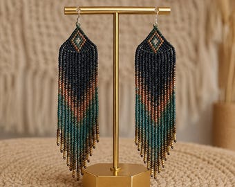 May include: A pair of long, dangling earrings featuring a chevron pattern made of tiny seed beads. The beads transition in color from dark navy at the top to copper, teal, and gold at the bottom, ending in a fringe of individual bead strands.