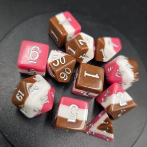 May include: A set of ten polyhedral dice featuring a tri-color design of brown, white, and pink horizontal stripes. Each die has silver-colored numbers engraved on the surfaces, including standard d20, d12, d10, d8, d6, and d4 shapes for gaming.