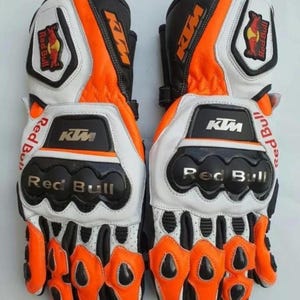 Puede incluir: Guantes de moto en blanco, negro y naranja. Presentan los logotipos de KTM y Red Bull. El diseño incluye acolchado protector y ventilación, enfocados en la seguridad y el rendimiento para los motociclistas.