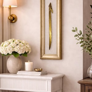 May include: A gold-framed wall art piece featuring a vertical, abstract gold design on a neutral linen background. The artwork hangs above a white table with a vase of white flowers, a candle, and a small gold bowl. A wall sconce provides soft lighting.
