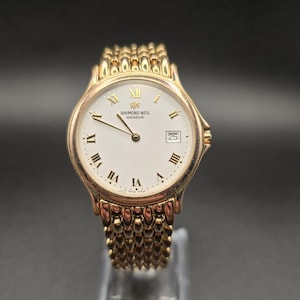 Può includere: Orologio Raymond Weil Geneve dorato con quadrante bianco, numeri romani e datario a ore 3. L'orologio ha un cinturino dorato e strutturato. L'orologio è esposto su un supporto trasparente.