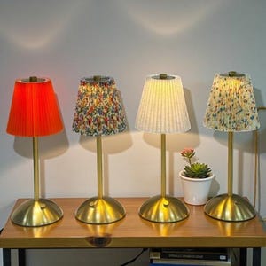 May include: Four decorative table lamps with gold bases and stems. The lampshades are pleated in various colours and patterns, including orange, floral, and cream. A small potted plant sits between two of the lamps on a wooden table.