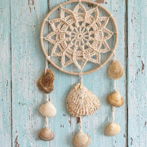 May include: A beige crochet dreamcatcher with a circular frame and a starburst design in the center. Shells of various sizes and colors hang from the bottom, suspended by twine. The background is a weathered, light blue wooden surface.