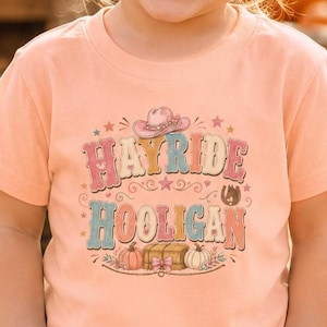 May include: A peach-colored short-sleeve t-shirt featuring a graphic print with the text "HAYRIDE HOOLIGAN" in a retro font, accompanied by illustrations of a cowboy hat, a horseshoe, and pumpkins, paired with light blue denim jeans.