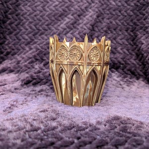May include: A gold-colored, decorative planter with an intricate Gothic architectural design. The planter features arched openings and floral rosette details, creating a vintage aesthetic. The planter is made of a rigid material and sits on a textured purple surface.