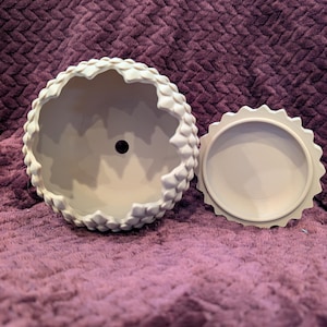 Könnte beinhalten: Ein hellbeige Keramik-Blumentopf mit einer strukturierten, schuppenartigen Außenseite und einem passenden Deckel. Der Topf hat eine runde Form mit einem gewellten Rand. Der Deckel ist kreisförmig mit einem ähnlichen gewellten Rand. Der Topf ist leer.