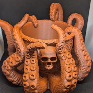 Könnte beinhalten: Ein orangefarbener, 3D-gedruckter, tintenfischförmiger Pflanzentopf mit einem Totenkopf. Die Tentakel umschließen eine zentrale Schale, mit dem Totenkopf an der Vorderseite. Der Topf hat detaillierte Texturen und einen dunklen Hintergrund.