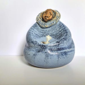 May include: A ceramic oil lamp shaped like a figure wearing a blue speckled parka with a hood. The figure has a brown face and a white wick protruding from the center of the blue body. The piece features a textured, mottled blue glaze finish.